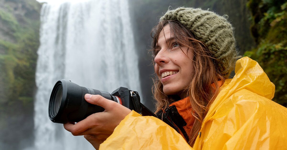 Woman with a raincoat on using a DSLR camera with a waterfall in the background.
