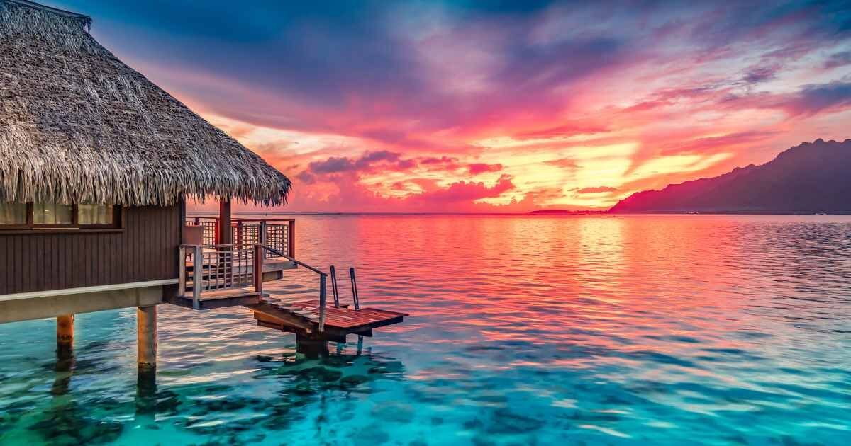 Overwater villa at sunset with vibrant pink, orange, and purple hues in the sky reflecting on the ocean.